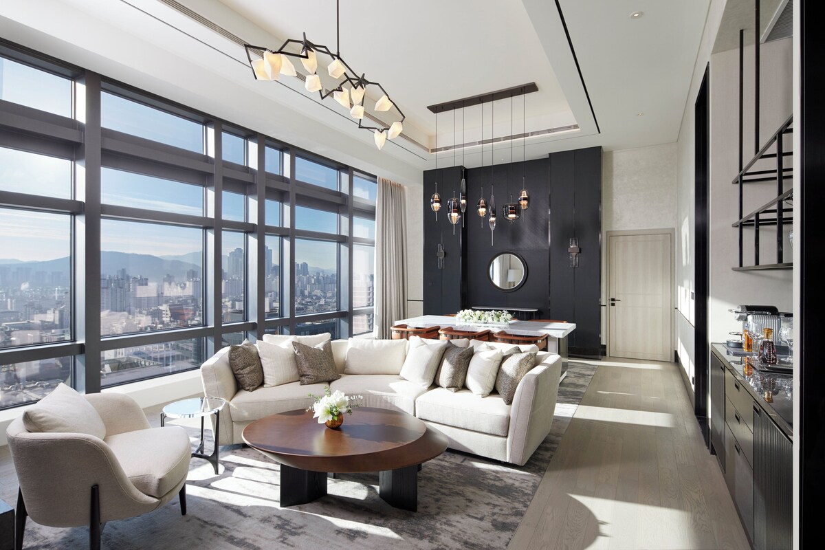 A modern living room with a large window, a sofa, a coffee table, and a kitchen area with a dining table.