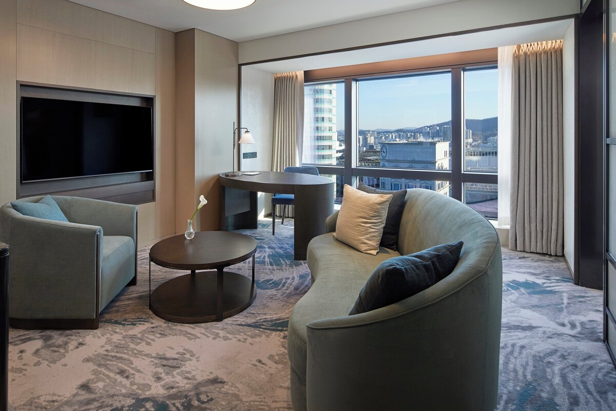 A modern hotel room with a large flat-screen TV, a sofa, a coffee table, and a dining area with a view of the cityscape.