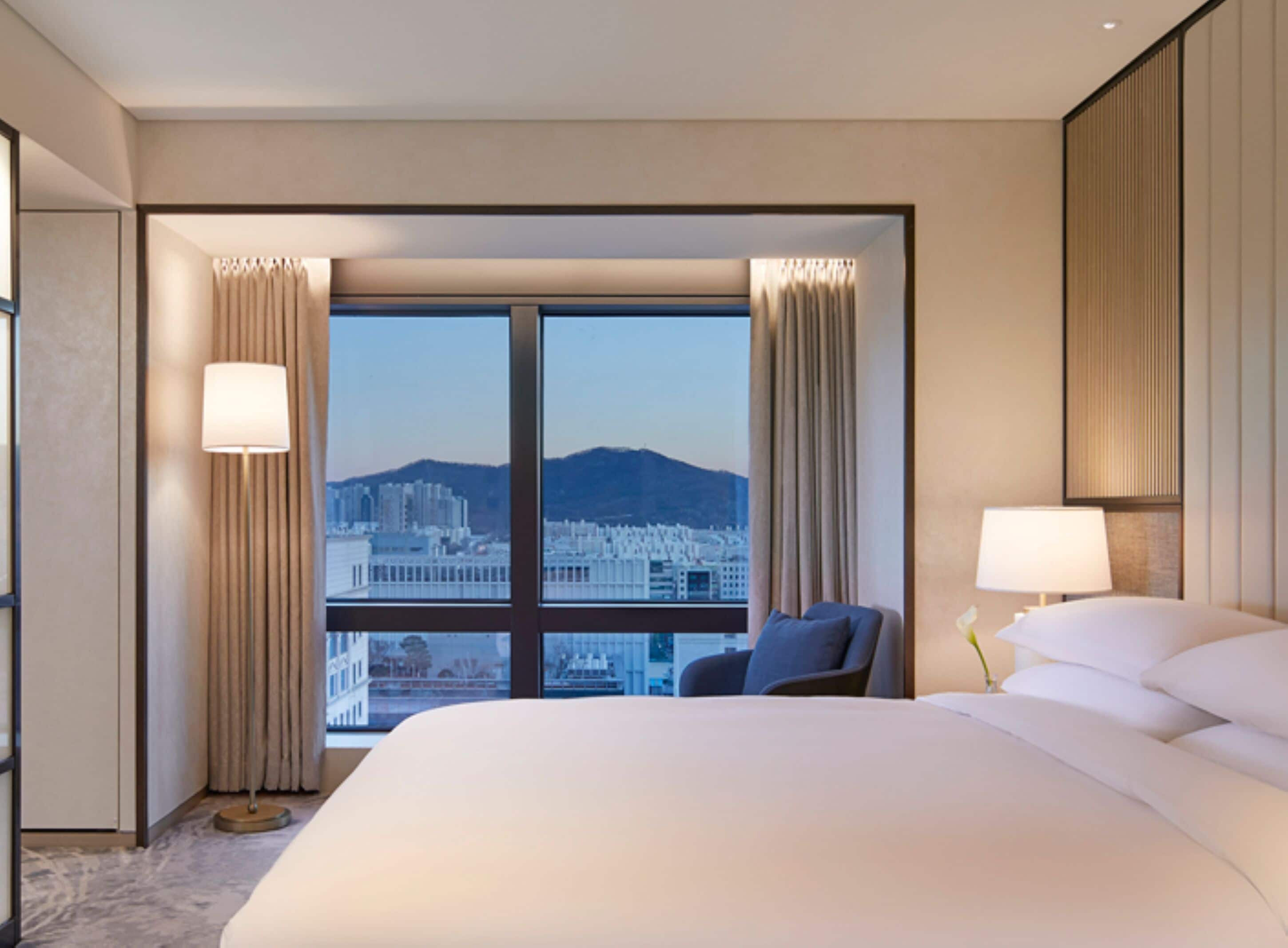 A hotel room with a large bed, a chair, a desk, and a view of a cityscape through large windows.