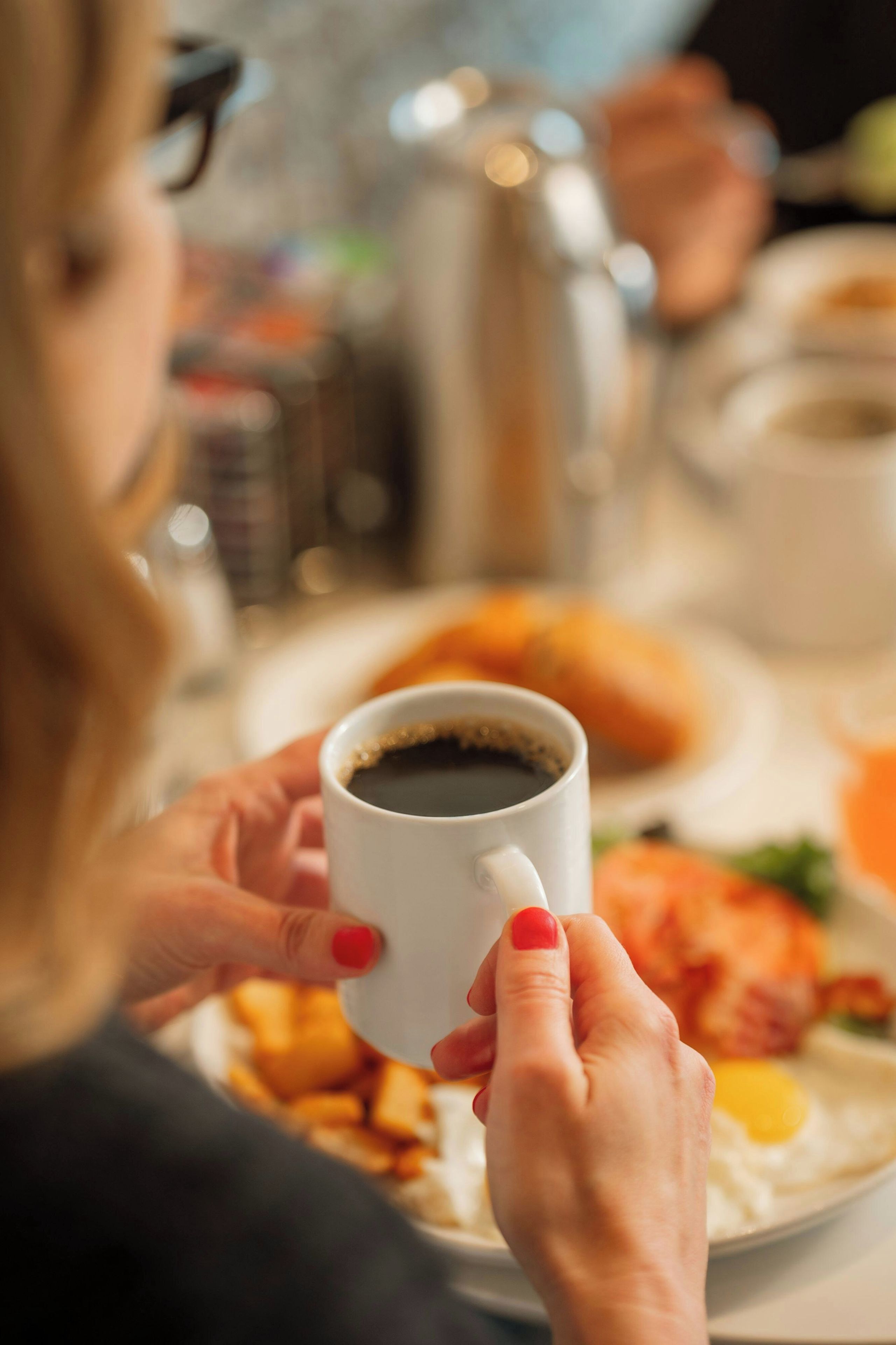 A person holding a cup of coffee, with a plate of food and a kettle in the background.