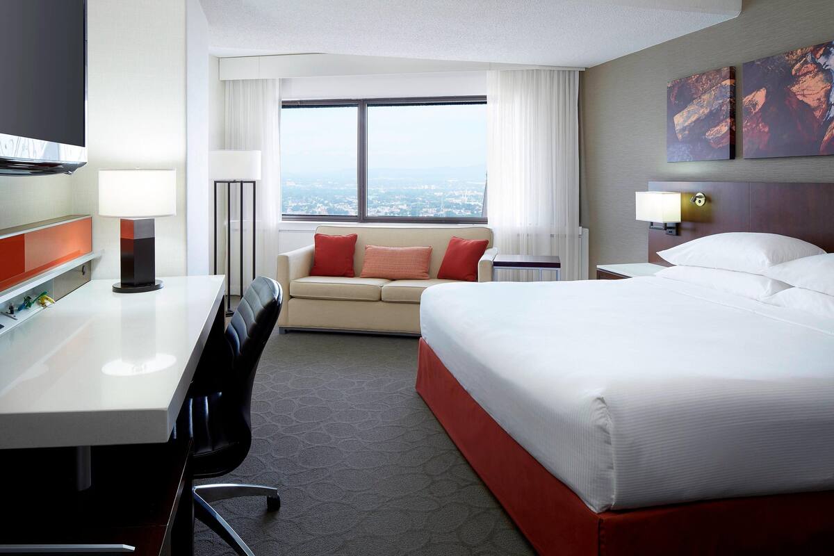 A hotel room with a bed, a desk, a sofa, a TV, and a large window with a city view.