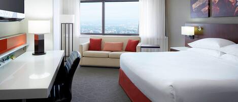 Ein Hotelzimmer mit Bett, Schreibtisch, Sofa, Fernseher und einem großen Fenster mit Blick auf die Stadt.