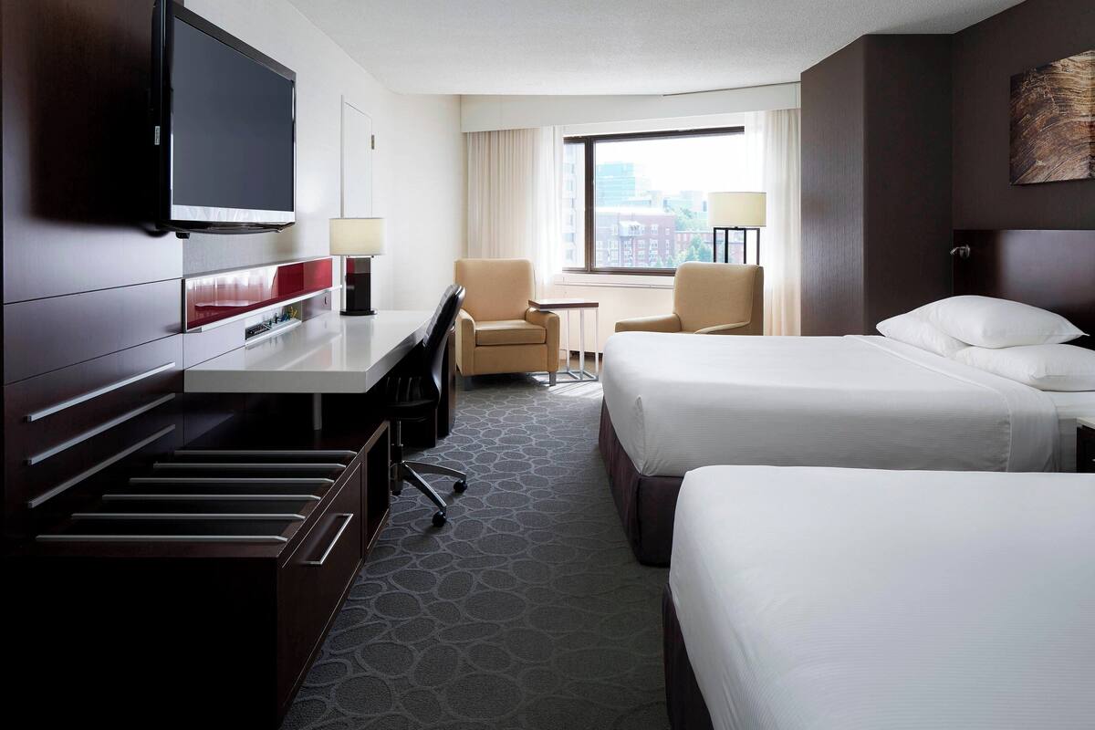 A hotel room with a flat-screen TV, a desk, two beds, a chair, and a window with a city view.