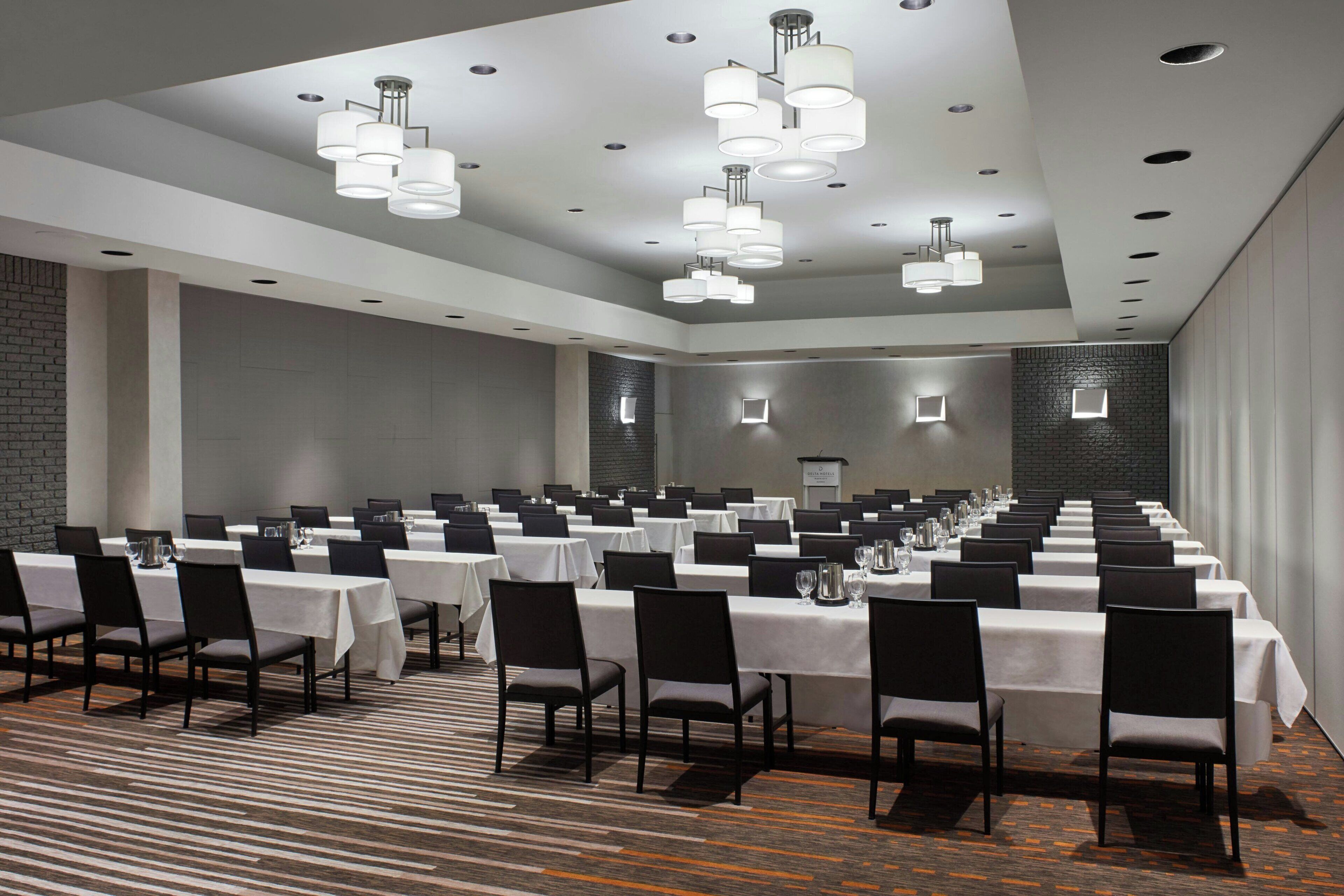 A conference room with multiple tables and chairs, arranged in rows, with overhead lighting and a patterned carpet.