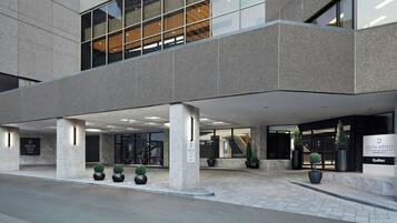 Una entrada de hotel moderna con un área exterior techada, ventanas amplias y plantas en macetas.