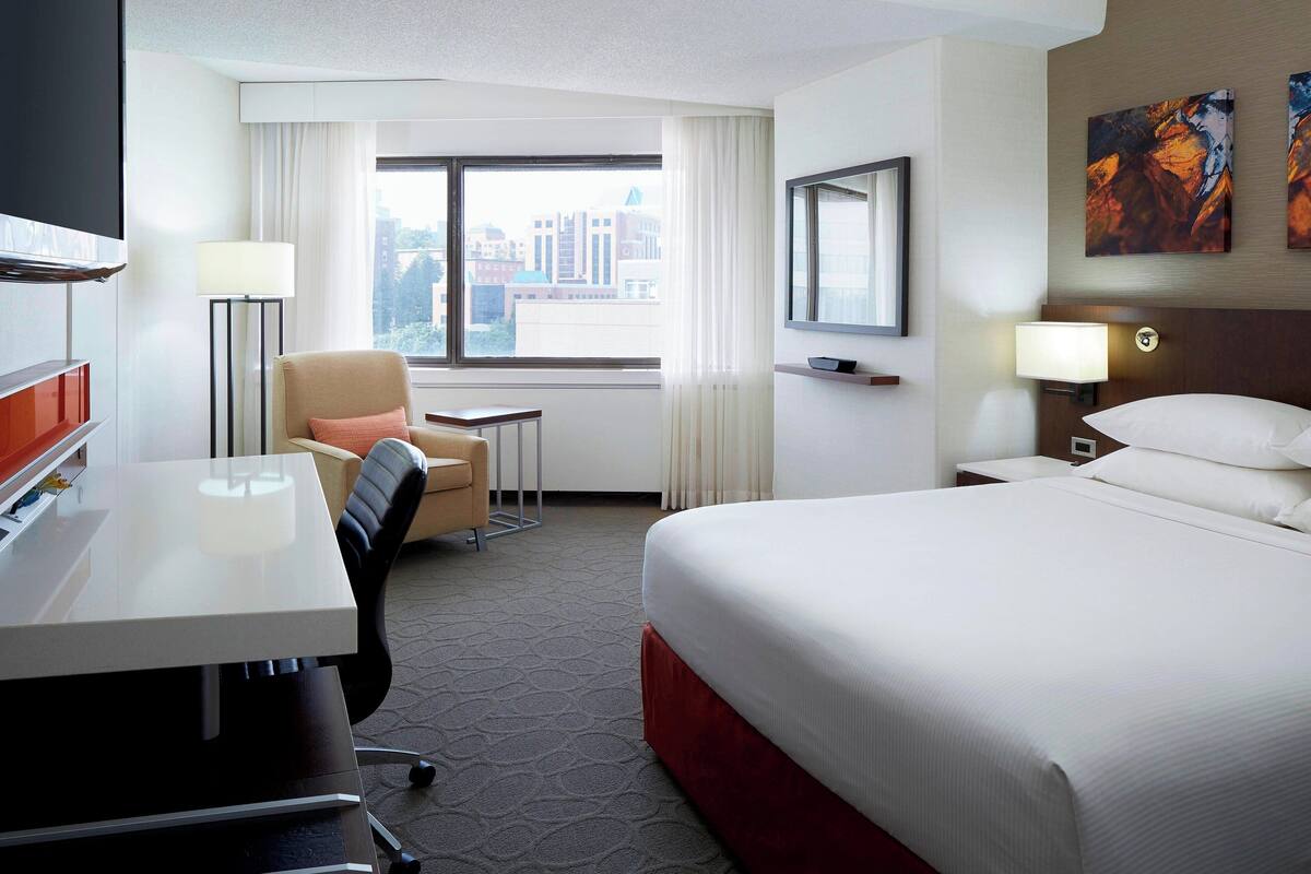 A hotel room with a bed, desk, chair, and a window with a city view.