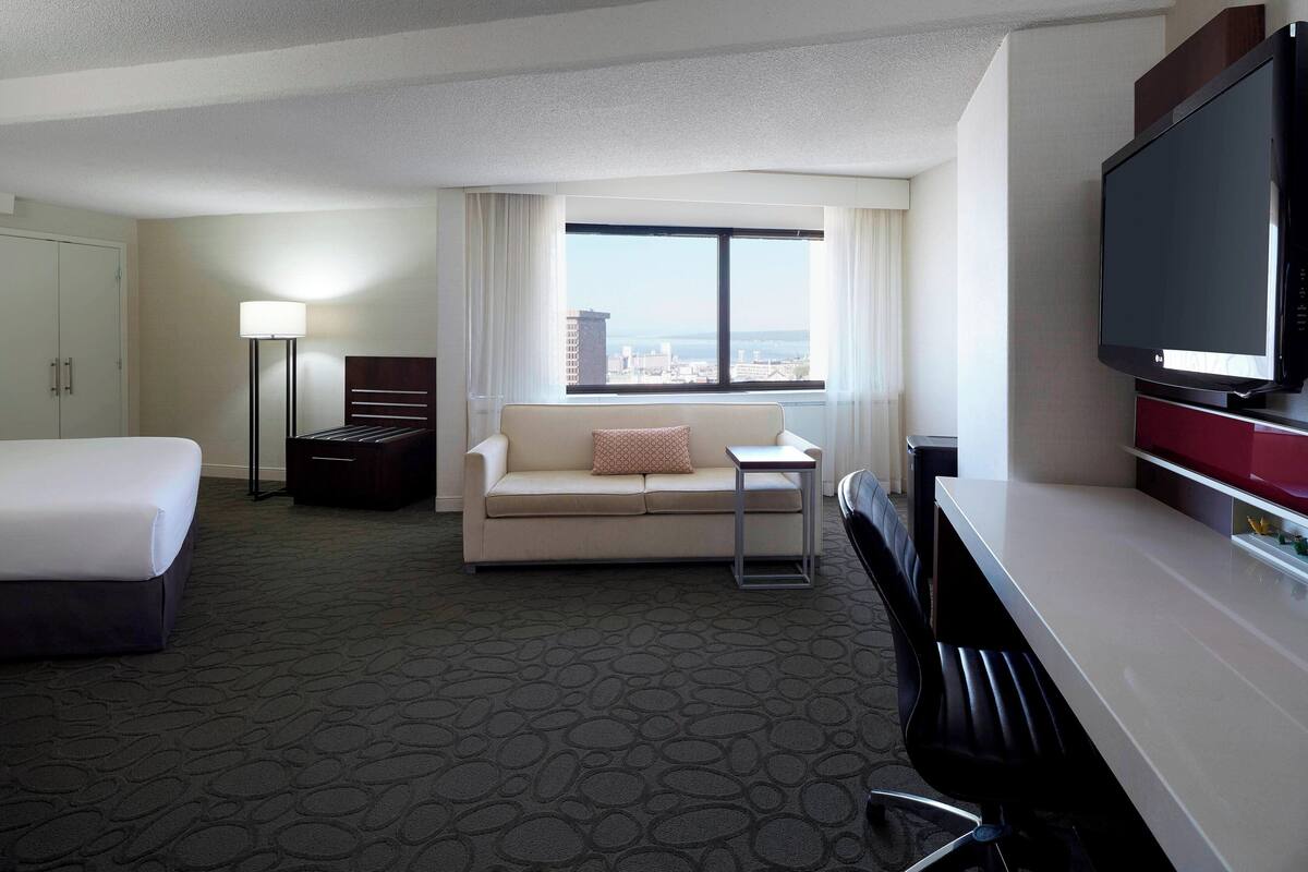A hotel room with a bed, a sofa, a desk, a TV, and a window with a city view.