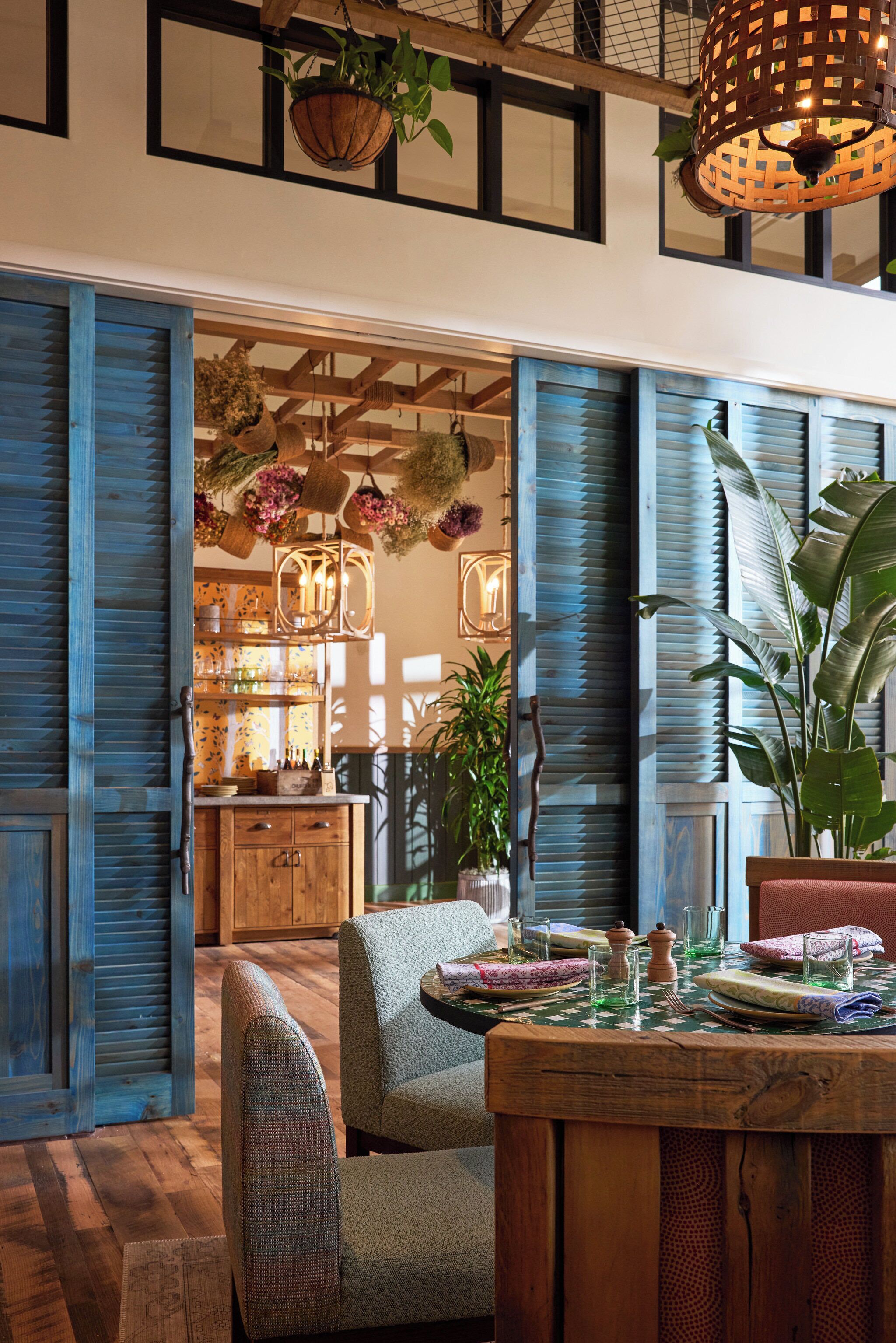 A dining area with a wooden table set for two, surrounded by blue shutters and hanging plants.