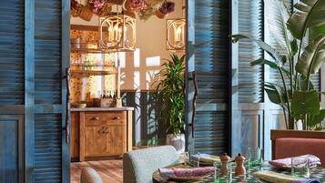 A dining area with a wooden table set for two, surrounded by blue shutters and hanging plants.