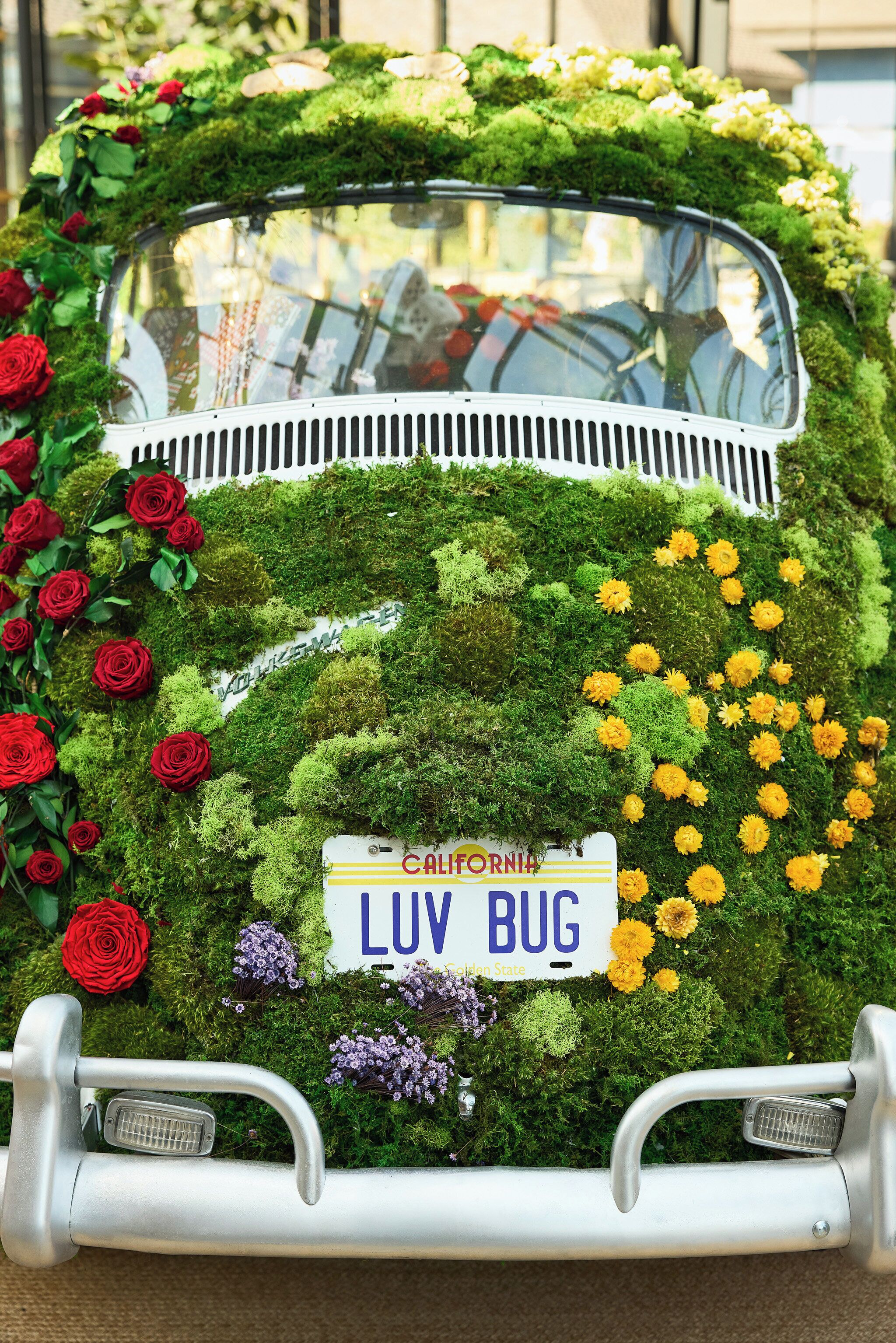 Un vehículo cubierto de musgo verde y flores, con una placa de California que dice 'LUV BUG'.