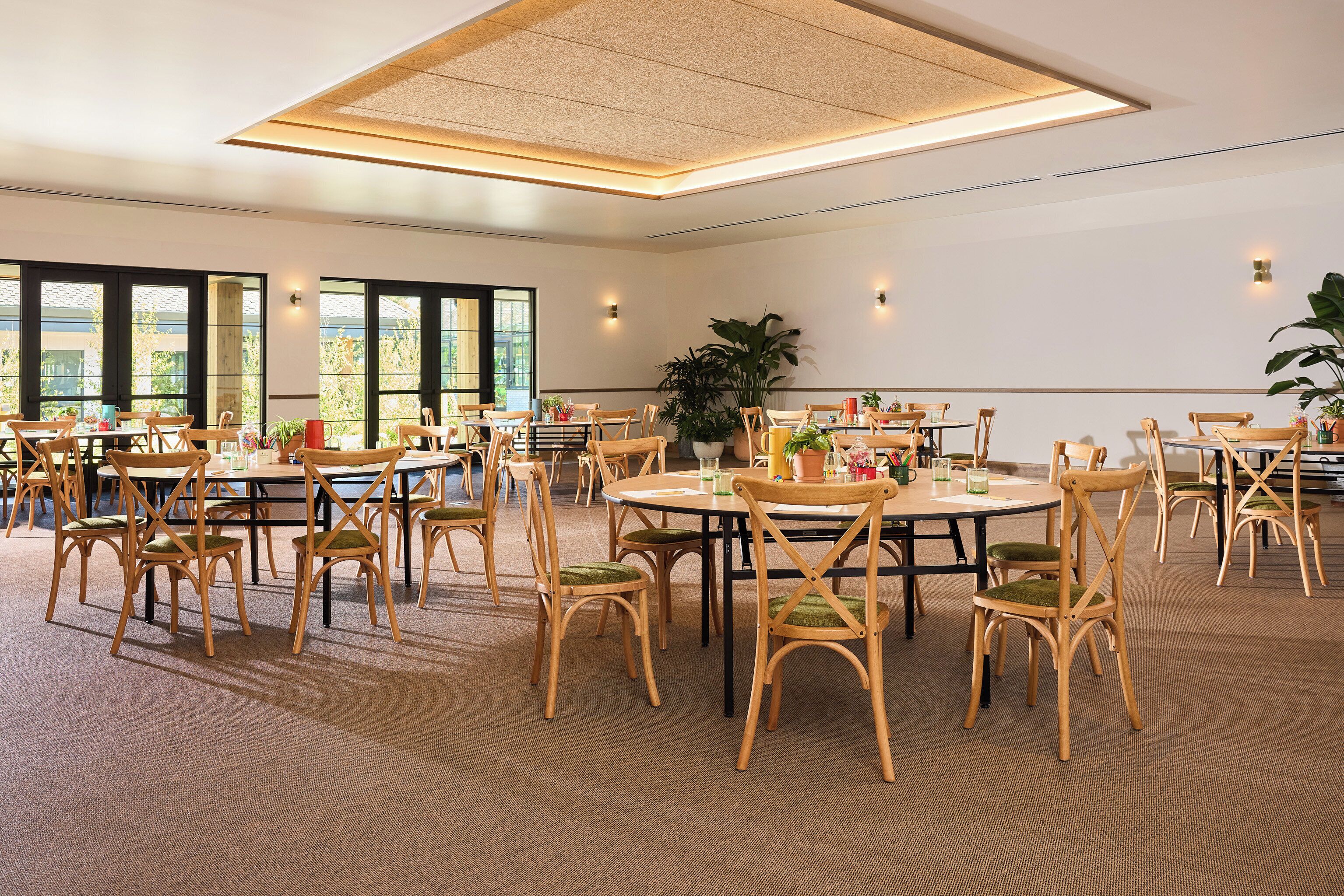 A spacious conference room with multiple round tables and chairs, large windows, and a ceiling light fixture.