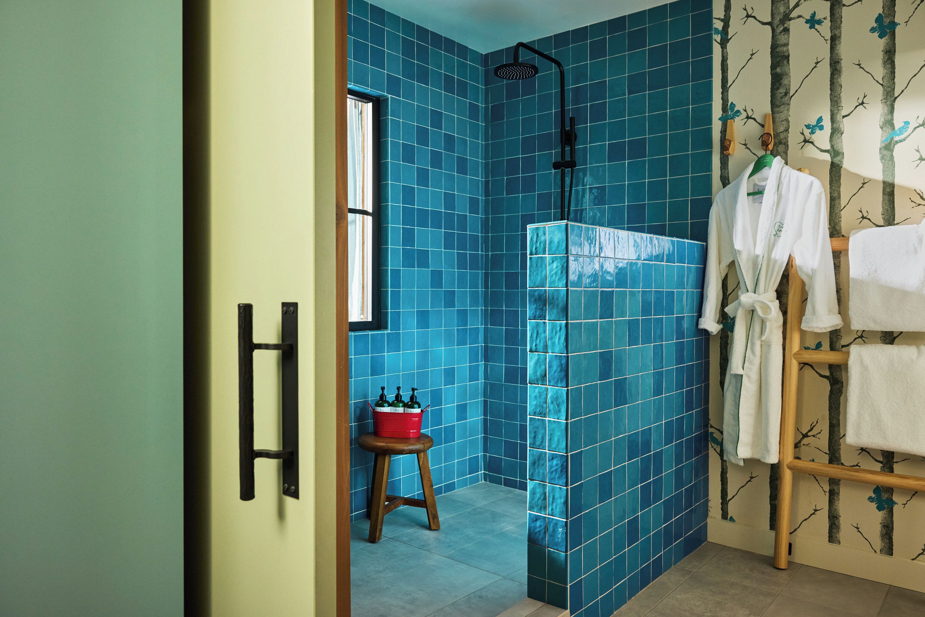 A bathroom with blue tiles, a shower, a wooden stool with a red bucket, and a towel rack with white towels.