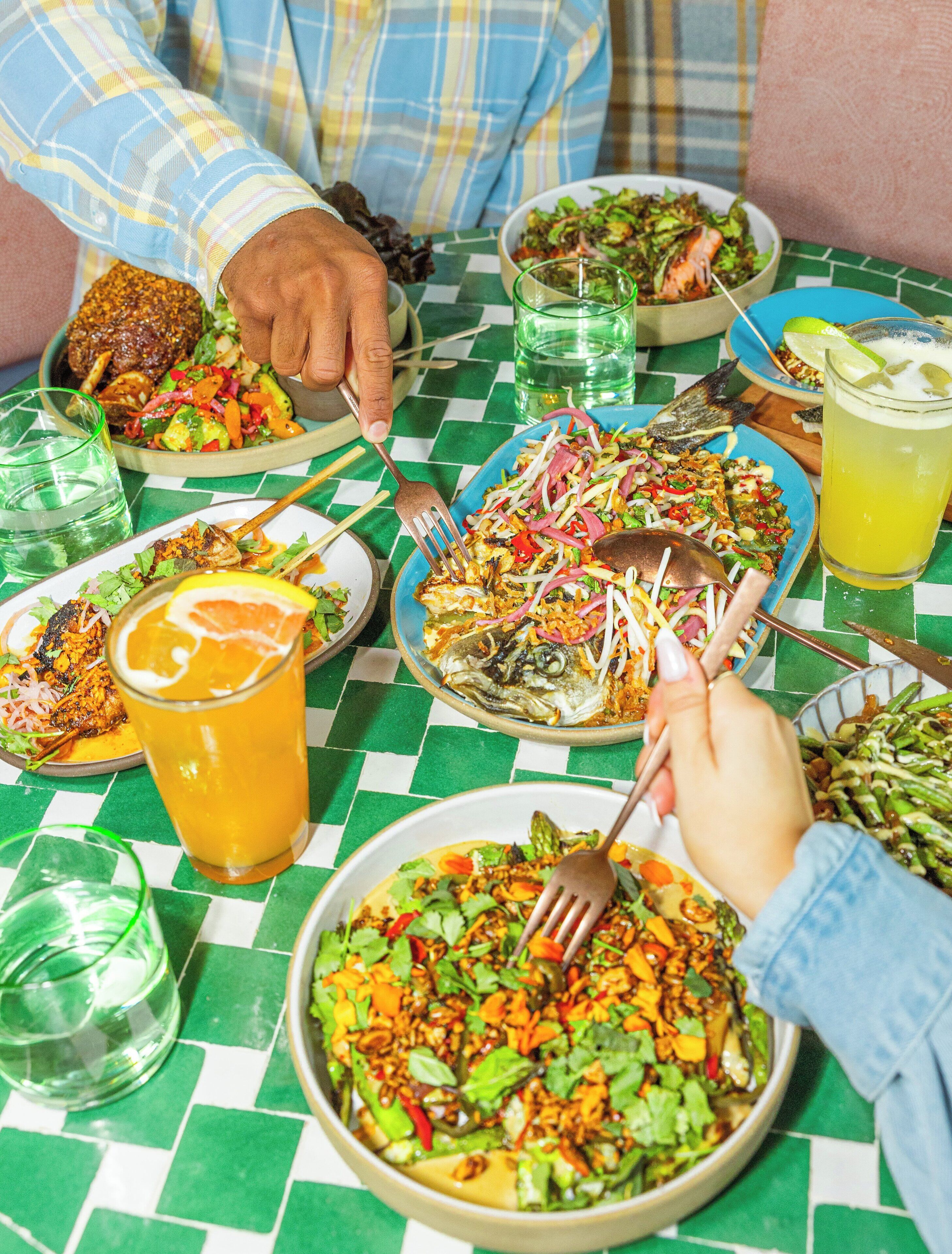 Une table à manger avec différents plats, notamment du poisson grillé, des salades et un bol de nouilles. On y voit des verres remplis de boissons et une personne qui utilise des baguettes.