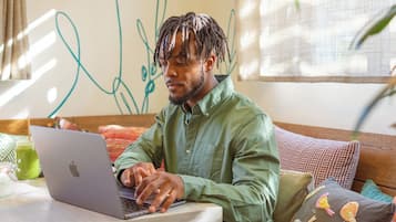A person sitting on a sofa with a laptop, a table with a drink, and a wall with abstract drawings.