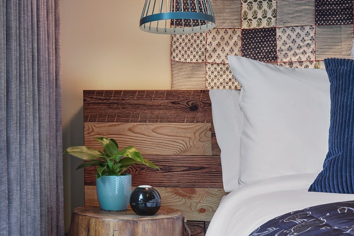 A bedroom with a wooden headboard, a blue and white patterned bedspread, a small wooden stool with a heart design, a potted plant, and a hanging light fixture.