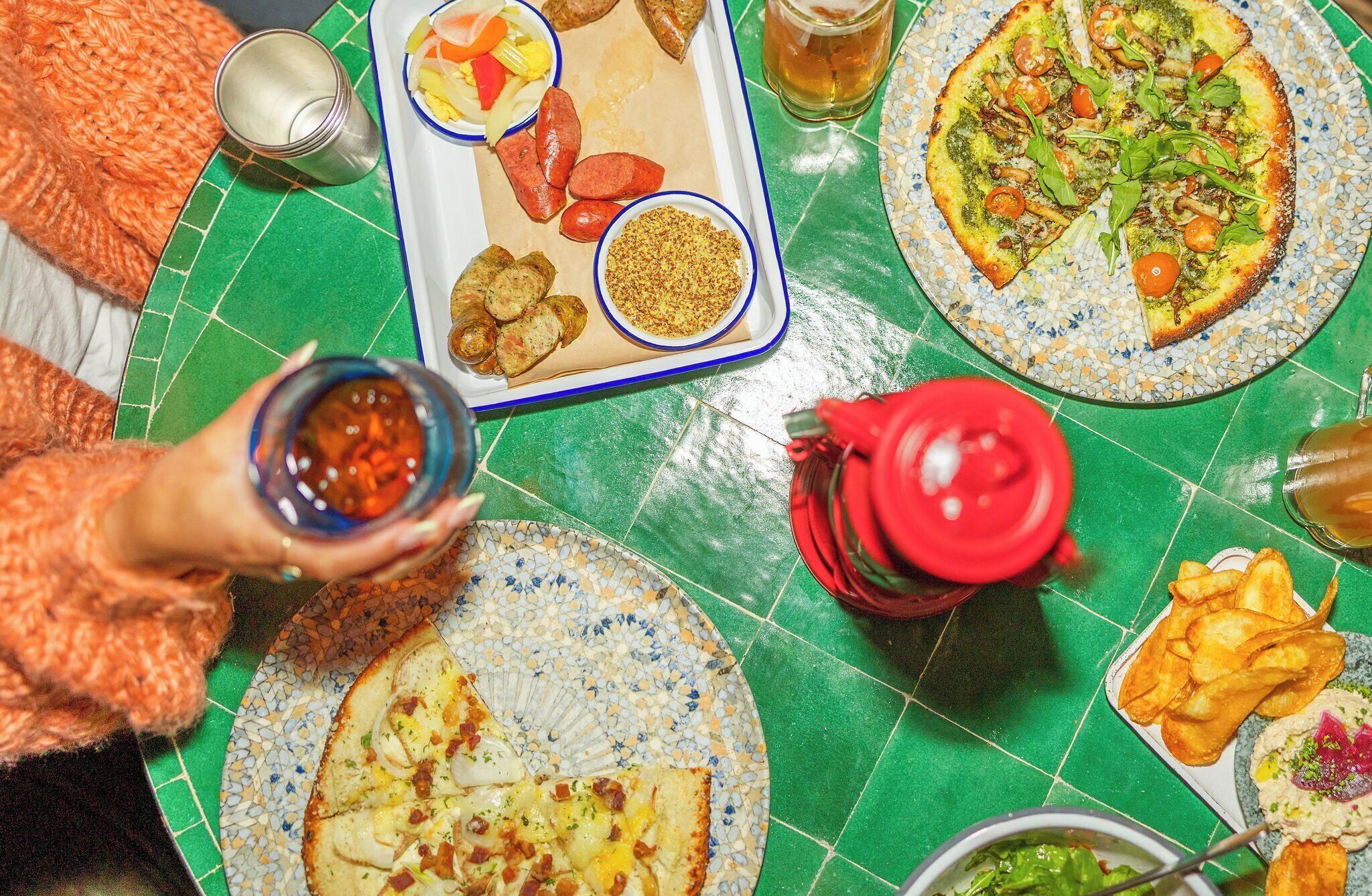 Una mesa de comedor con diversos platos, entre ellos pizzas y una bandeja con salchichas y verduras.