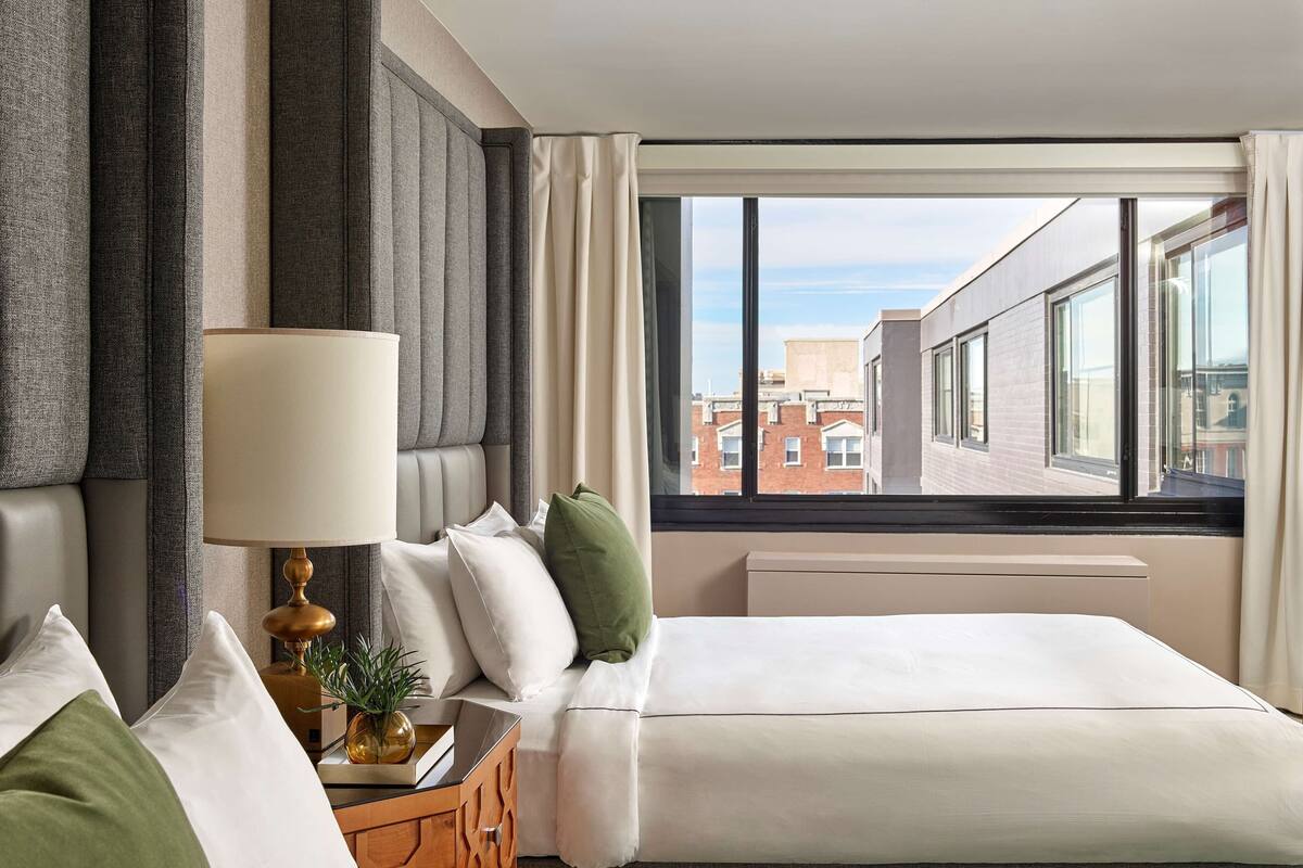 Habitación de hotel con dos camas, una mesita de noche con lámpara y vistas a la ciudad a través de amplios ventanales.