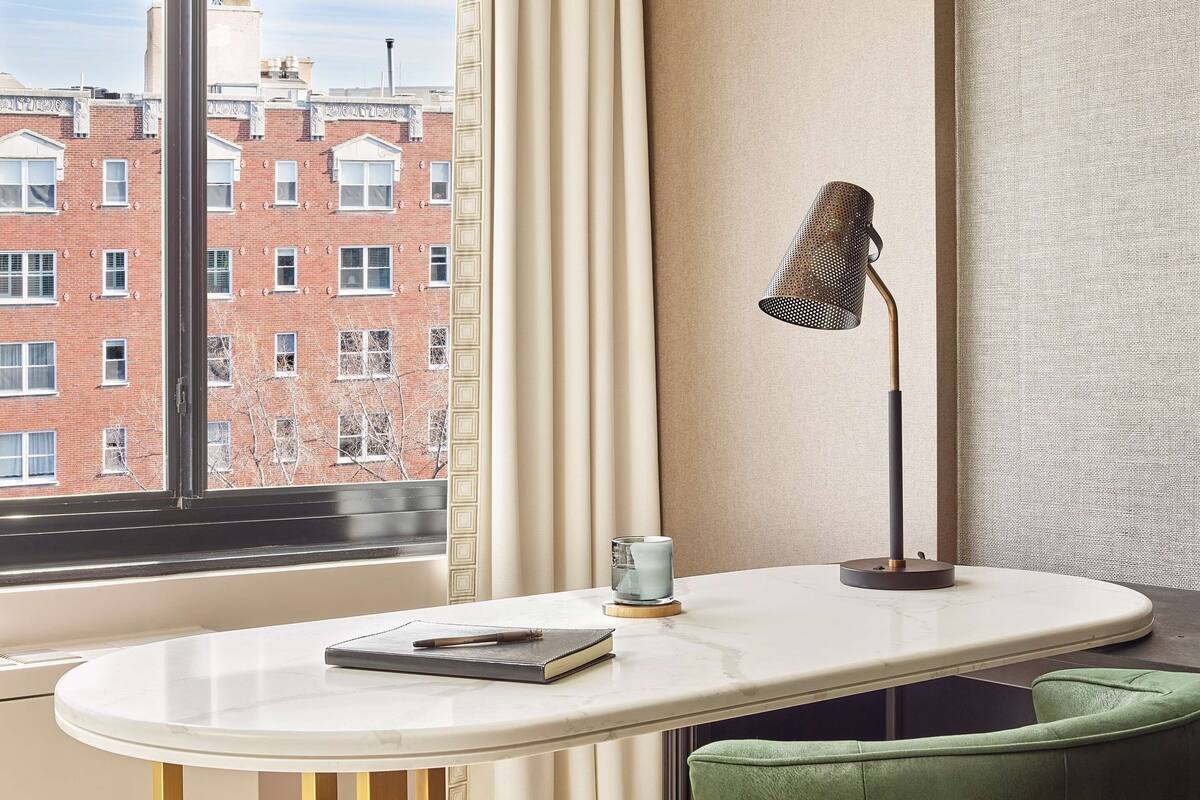 Habitación moderna con una mesa redonda, sillas tapizadas en verde, una lámpara y vista a un edificio de ladrillo a través de una ventana.