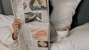 Eine Person liegt im Bett und liest eine Zeitung, mit einer Kaffeetasse und einem Croissant in der Nähe.