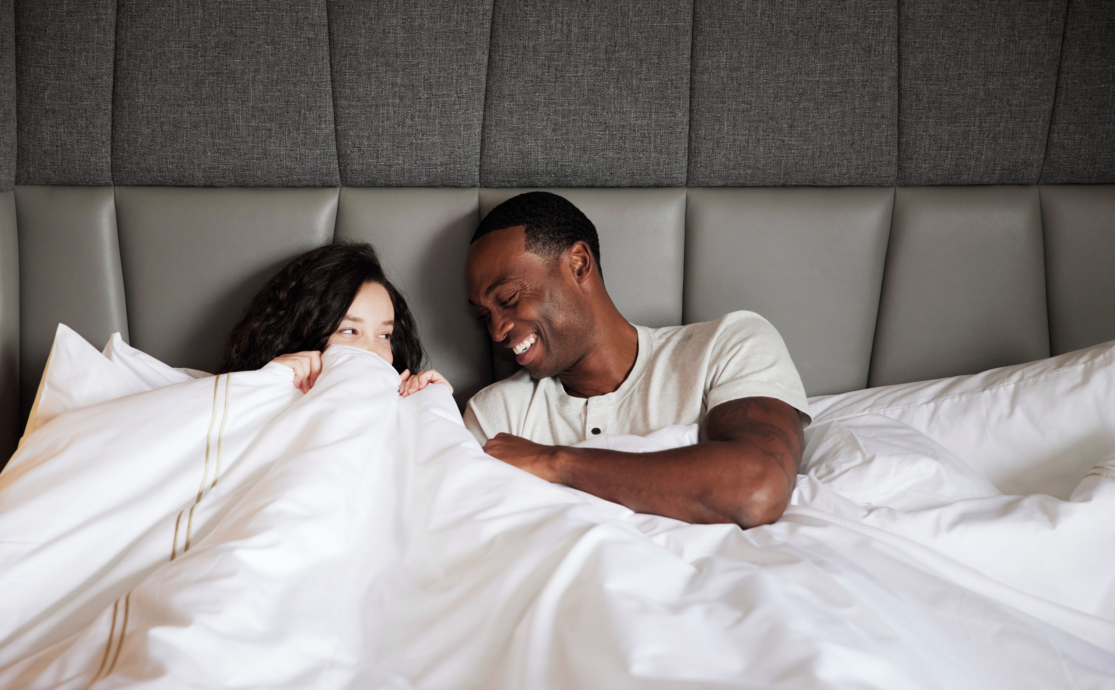 Une chambre à coucher avec une tête de lit, un lit recouvert de draps blancs, et une personne allongée sur le lit.