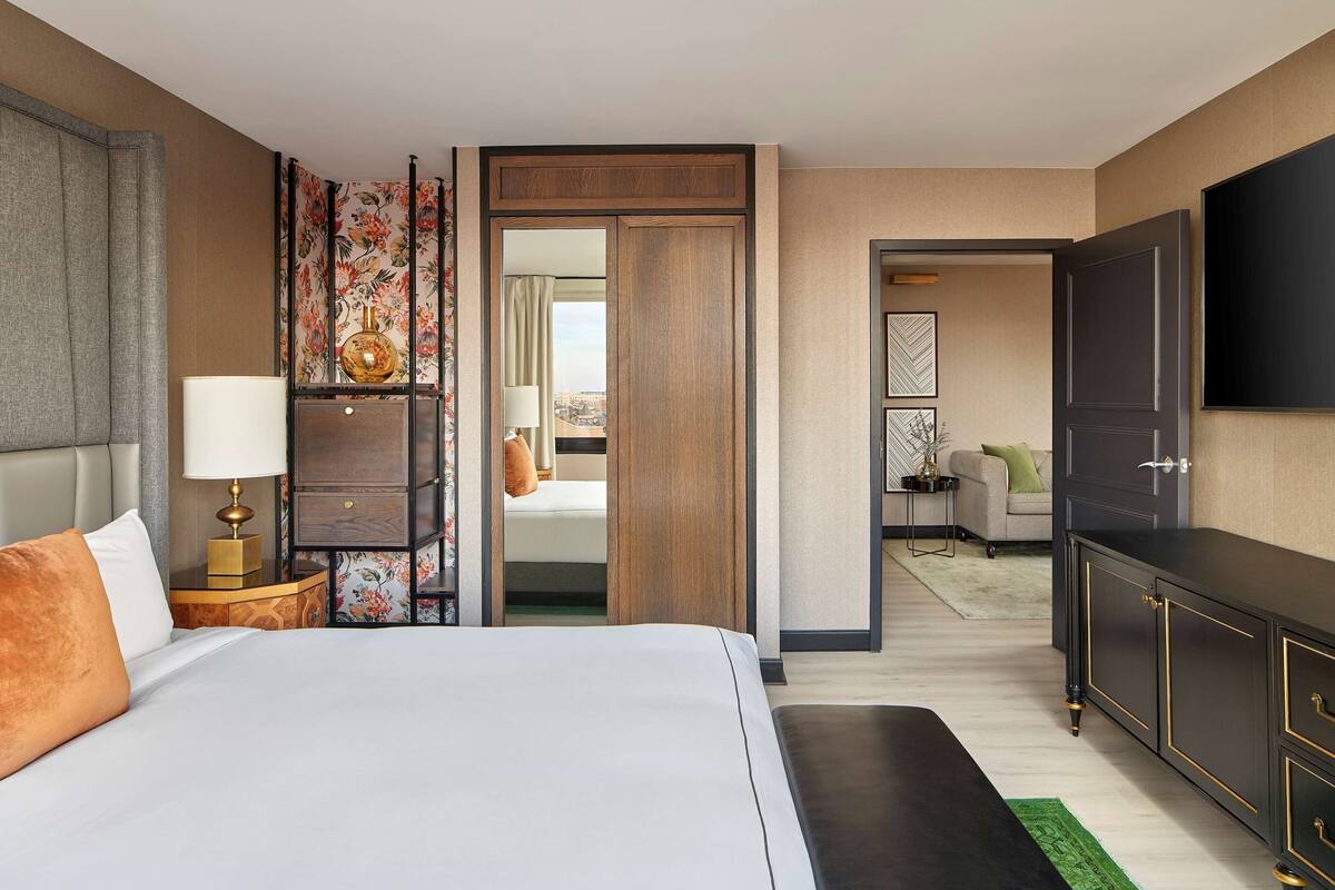 Una habitación de hotel moderna con una cama grande, una mesita de noche con lámpara, un armario y un televisor en la pared.