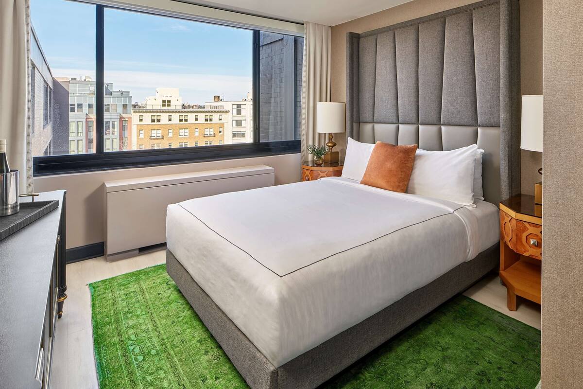 Habitación de hotel con una cama grande, vistas de edificios por la ventana, una mesita de noche con una lámpara y un radiador.
