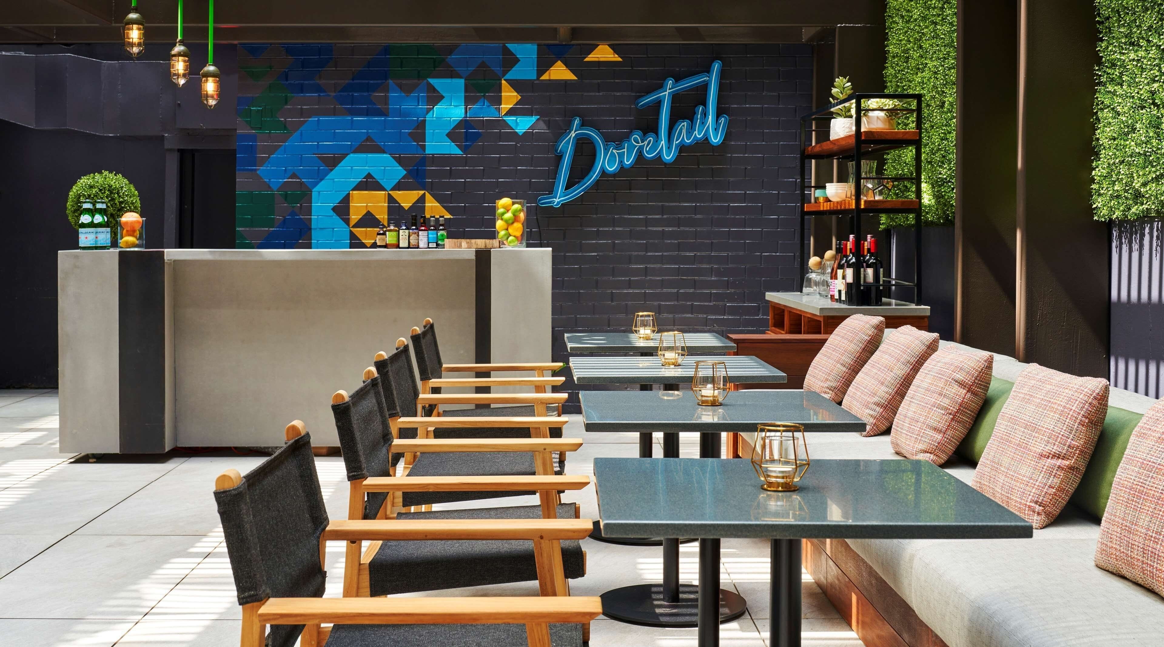 A modern bar area with a neon sign, wooden chairs, and tables, and a green wall with a geometric pattern.
