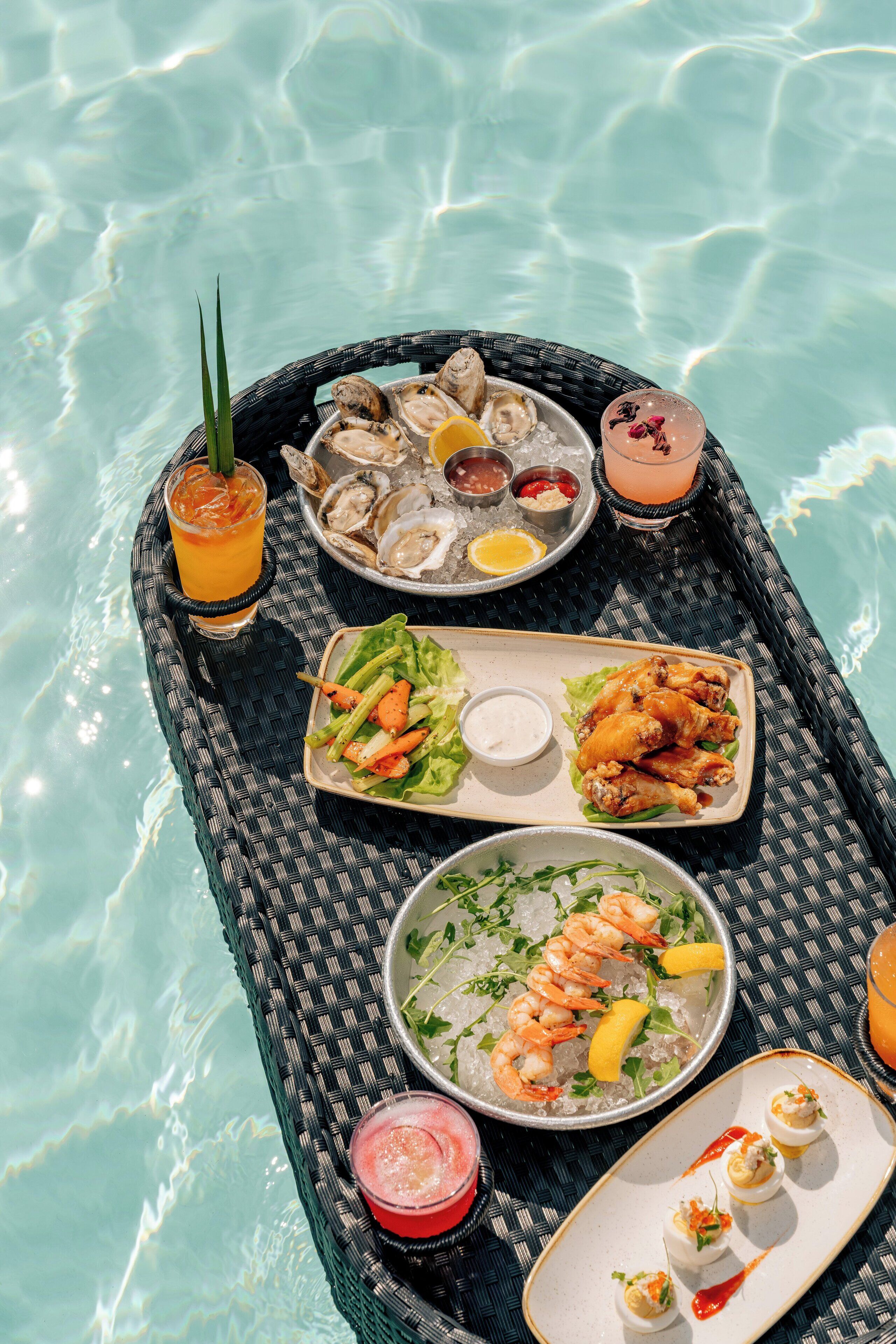 Un coin repas aménagé au bord d’une piscine, avec des plats de fruits de mer, des boissons et une table en osier.