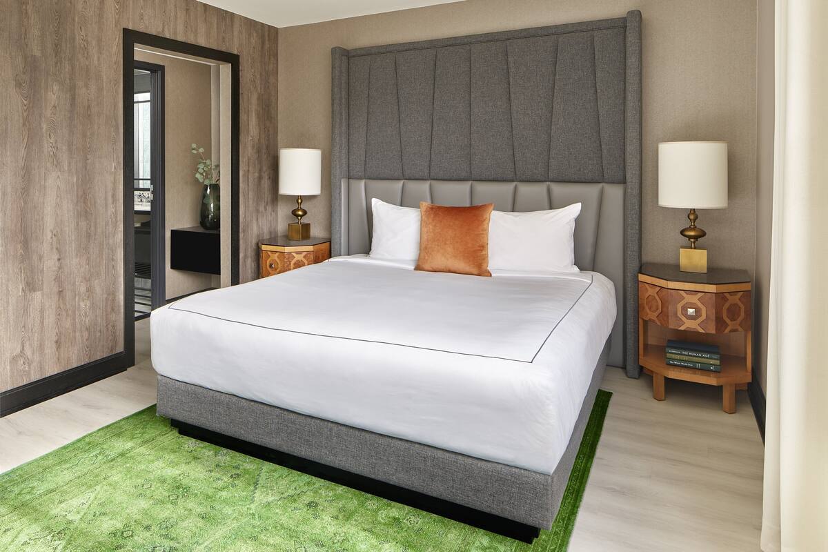 Una habitación de hotel moderna con una cama grande, cabecera tapizada en gris, dos mesitas de noche con lámparas y una alfombra verde.