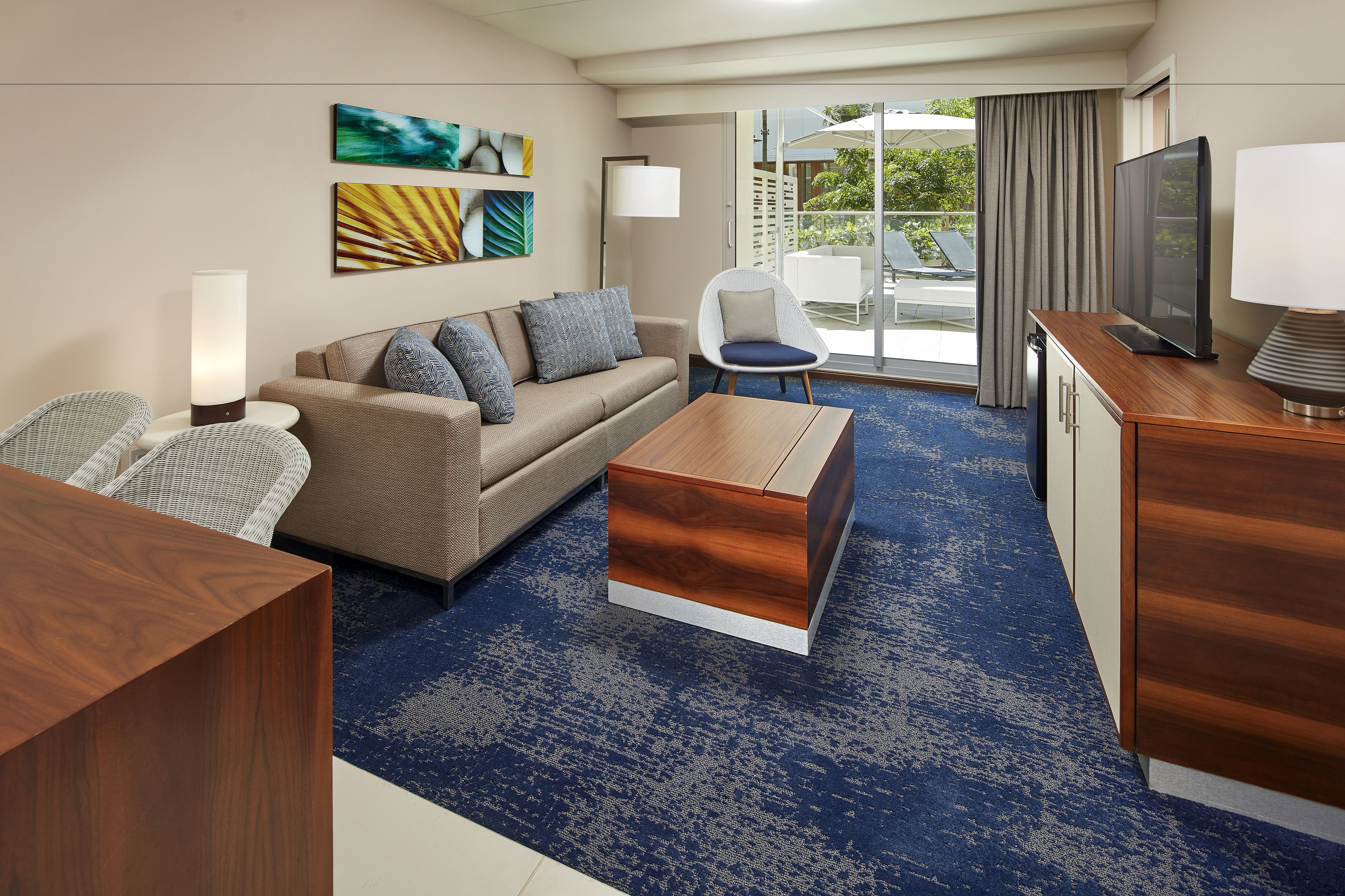 A modern hotel room with a sofa, armchair, wooden coffee table, flat-screen TV, and a view of the outdoor pool area.