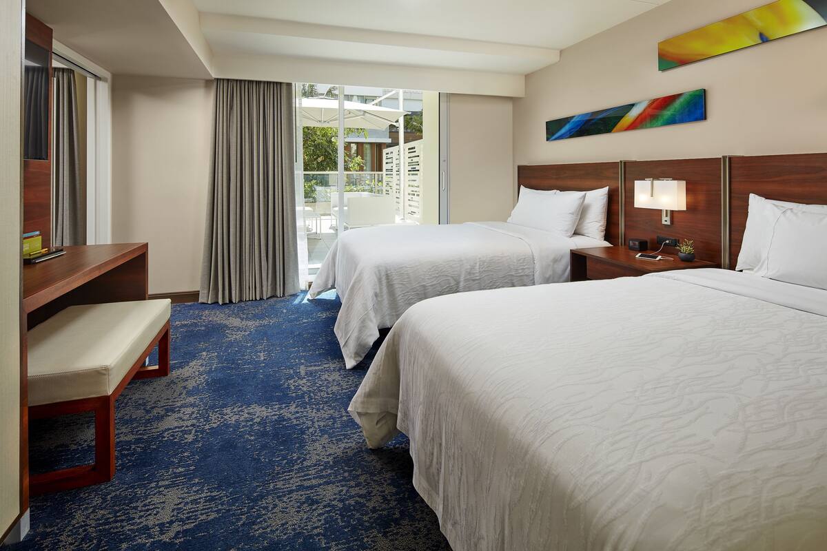 A hotel room with two beds, a bench, a desk, and a large window with curtains.