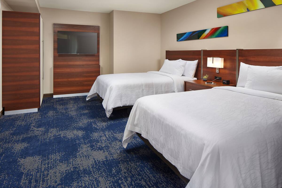 A hotel room with two beds, a flat-screen TV, a painting, and a nightstand with a lamp.