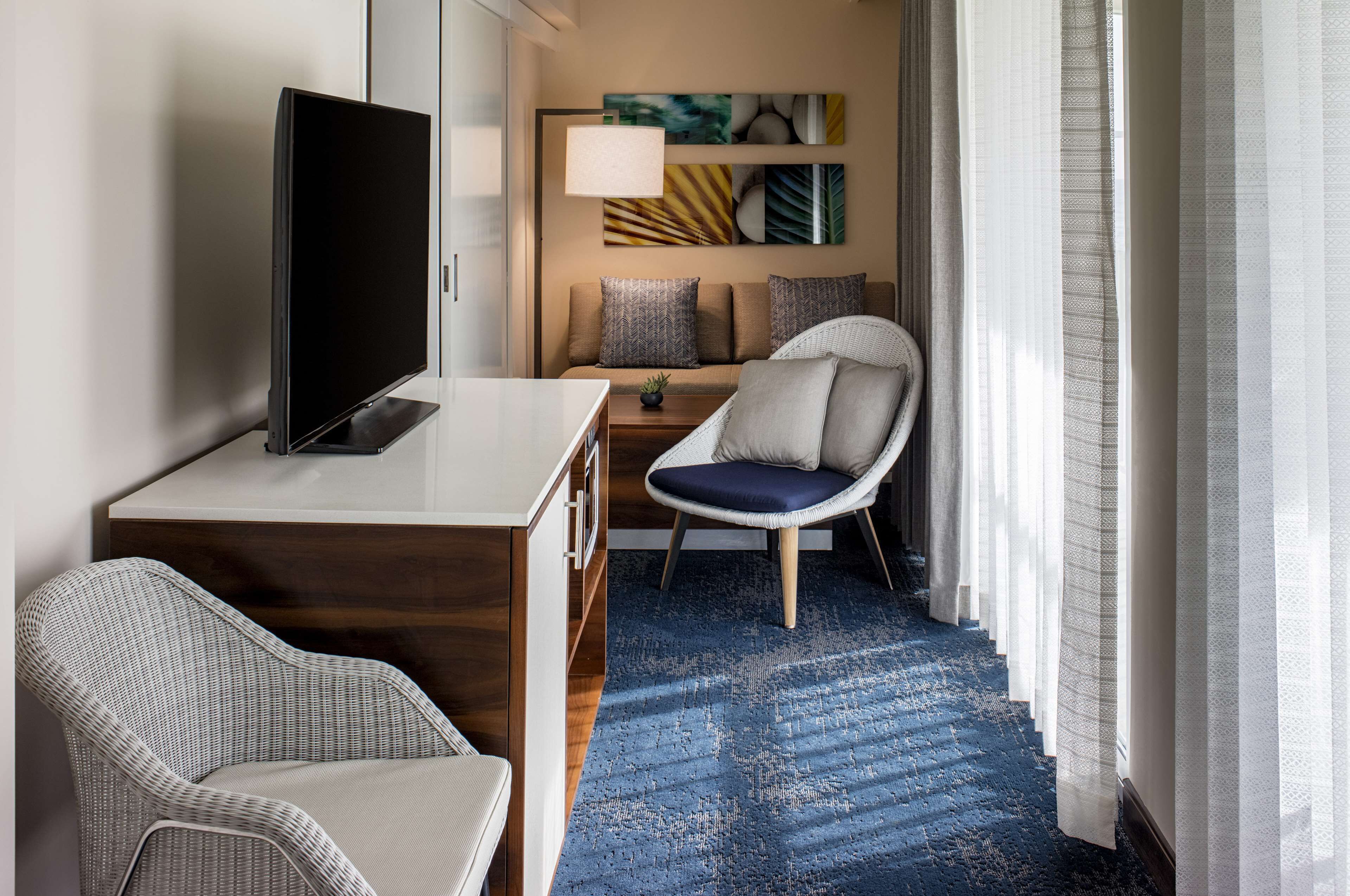 A hotel room with a flat-screen TV, a wicker chair, a sofa with patterned cushions, a small table with a lamp, and a blue patterned rug.