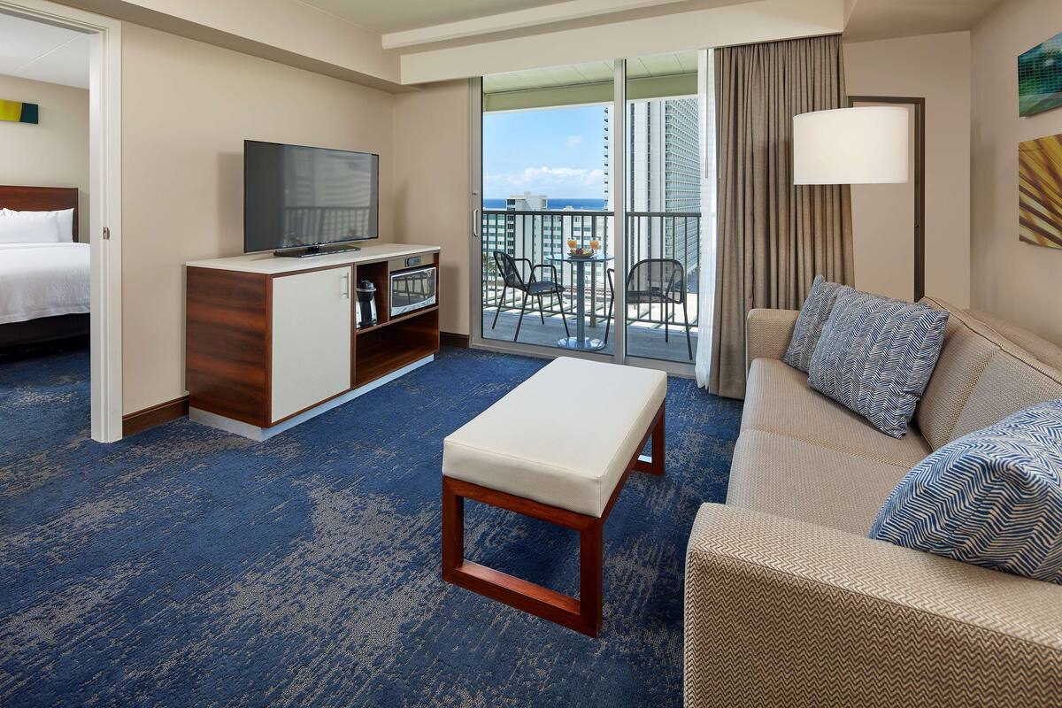 A hotel room with a balcony, a sofa, a coffee table, a TV, and a desk with a lamp.
