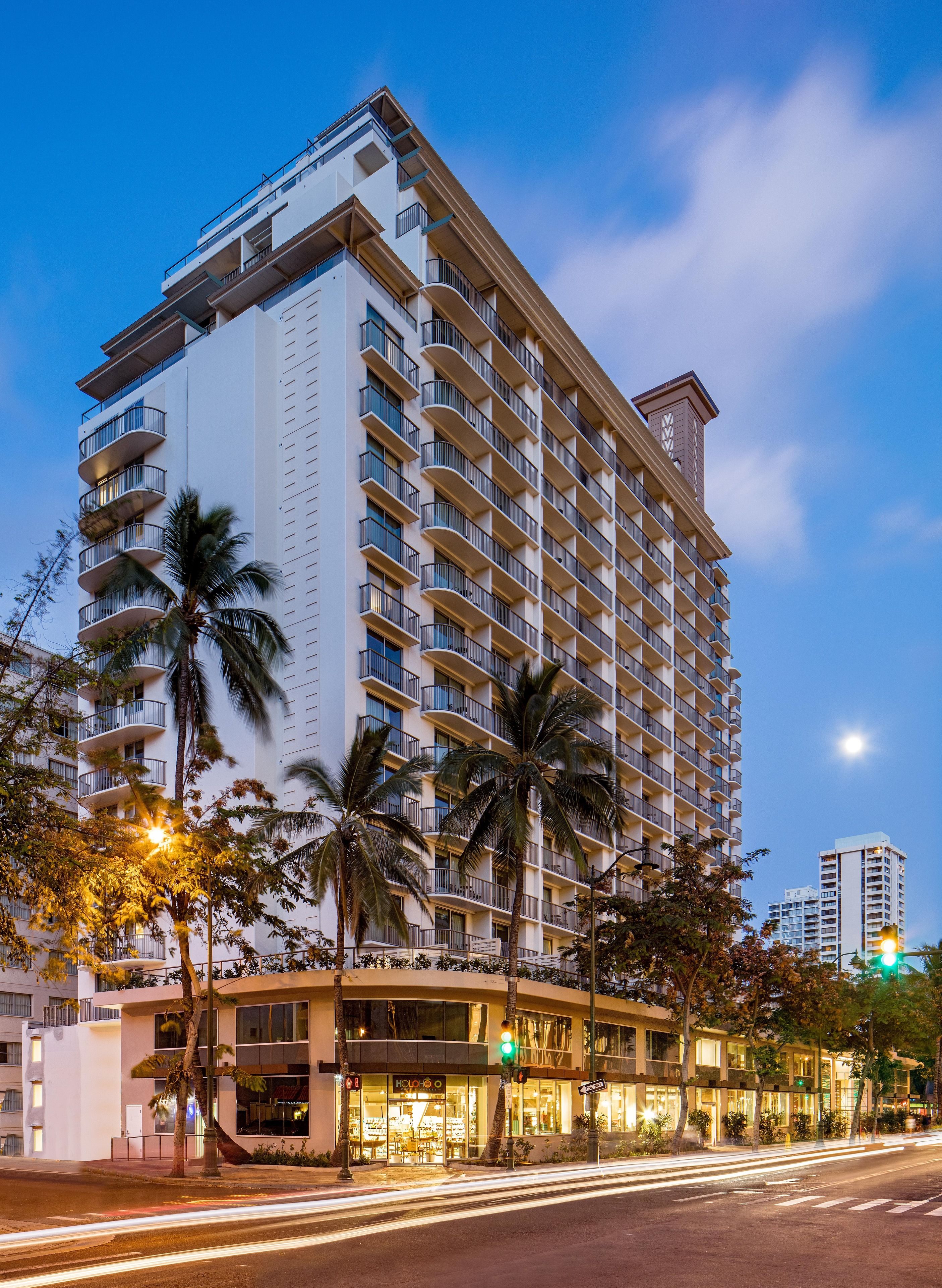 A high-rise hotel with multiple balconies, palm trees, and a storefront with illuminated signage.