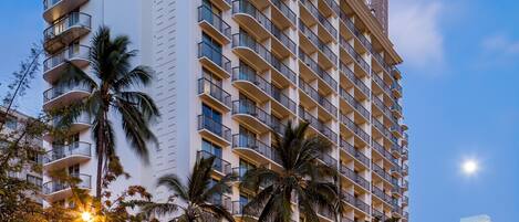 A high-rise hotel with multiple balconies, palm trees, and a storefront with illuminated signage.
