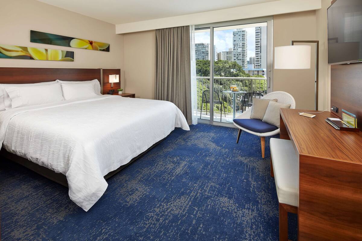 A hotel room with a large bed, a desk, a chair, a TV, and a balcony with a city view.