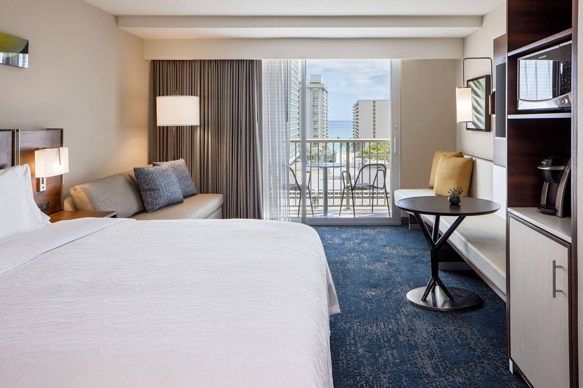 A hotel room with a large bed, a sitting area with a sofa and armchairs, a small round table, and a balcony with a view of the city.
