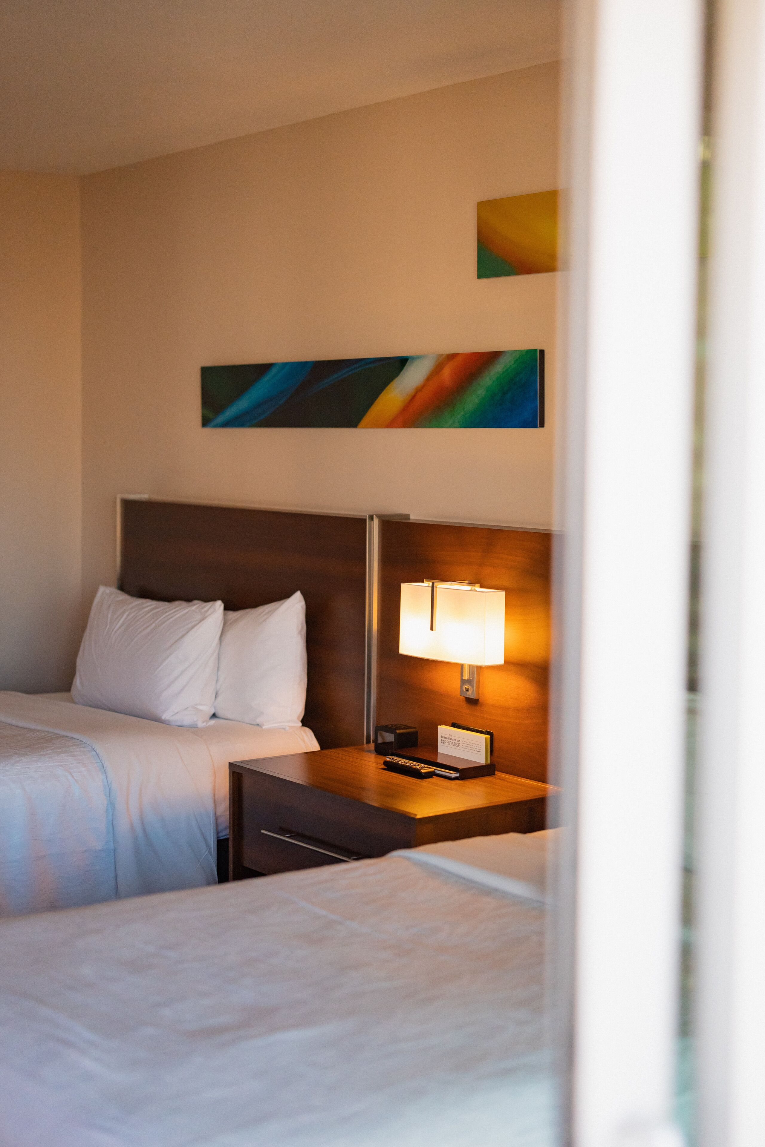 A hotel room with two beds, a nightstand with a lamp, and a painting on the wall.