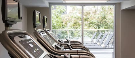 A row of stationary exercise bikes with digital displays, a large window with a view of outdoor seating, and a ceiling-mounted light fixture.