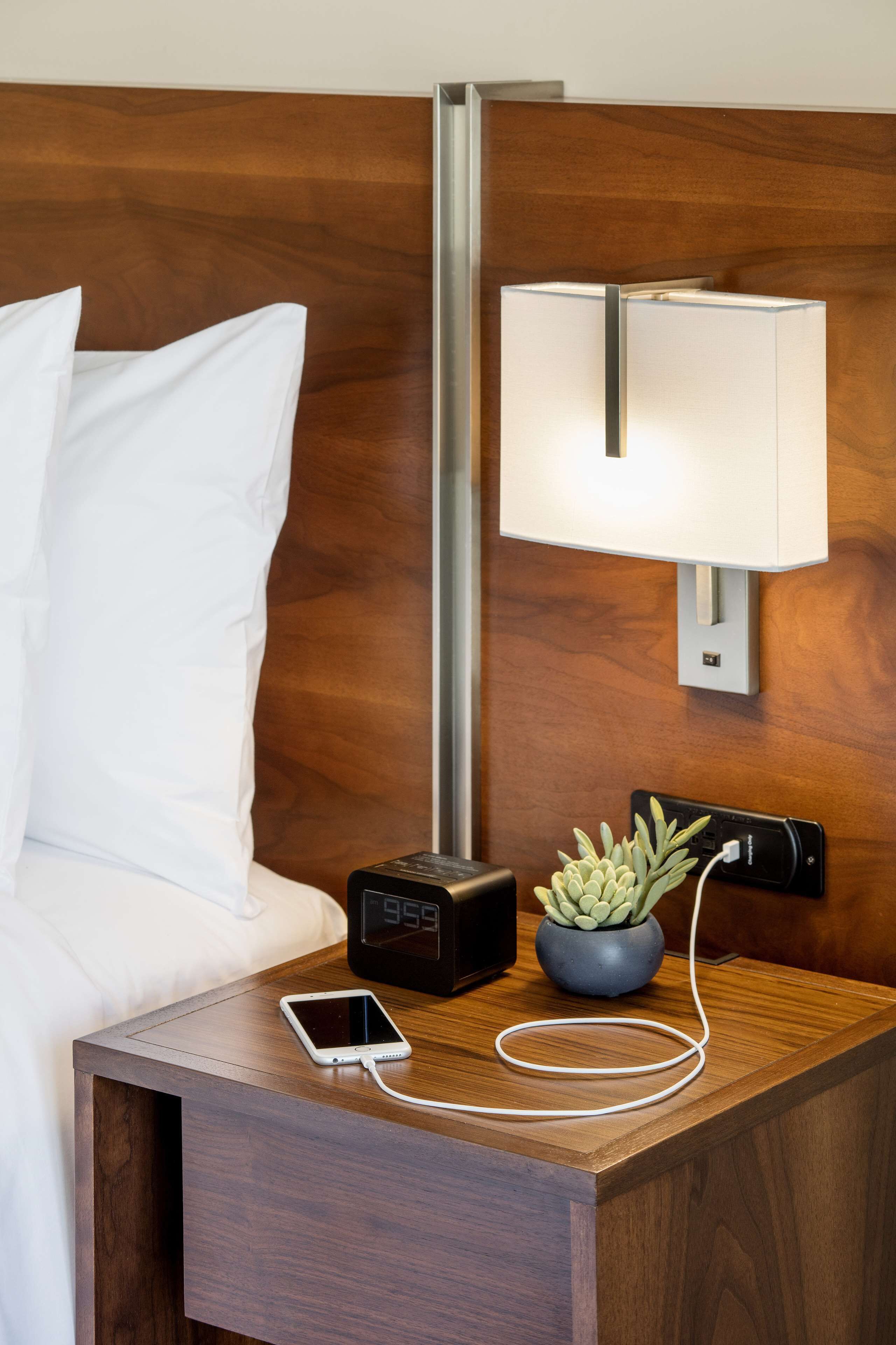 A neatly made bed with white linens, a bedside table with a digital clock, a smartphone, a small potted plant, and a wall-mounted lamp.