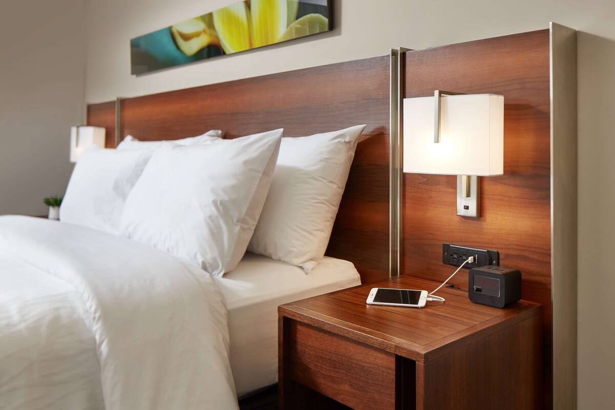 A neatly made bed with white linens, a wooden nightstand with a lamp, and a digital clock.
