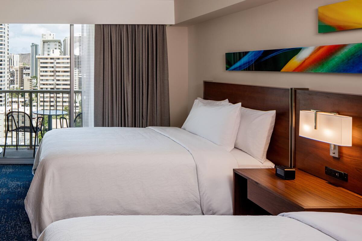 A hotel room with two beds, a view of the cityscape, a balcony with chairs, and a modern painting on the wall.