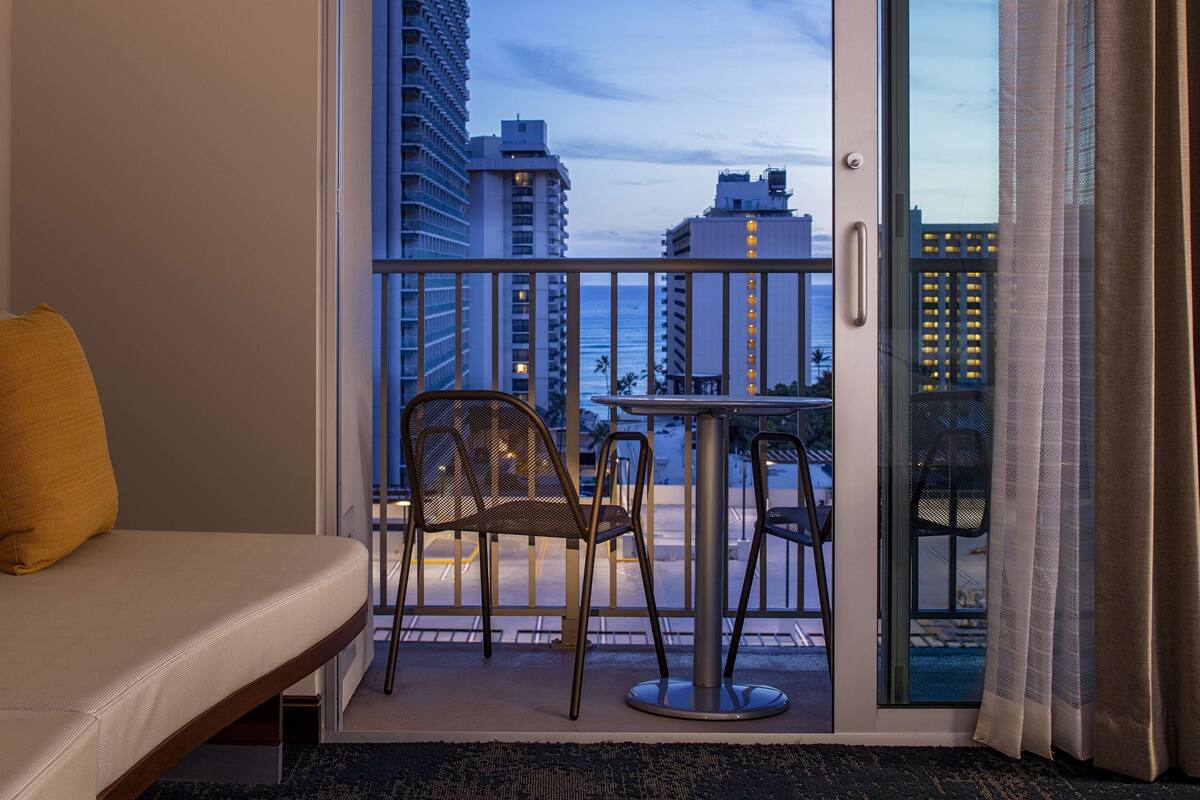 A balcony with a sofa, chairs, and a table, overlooking a cityscape and the ocean.
