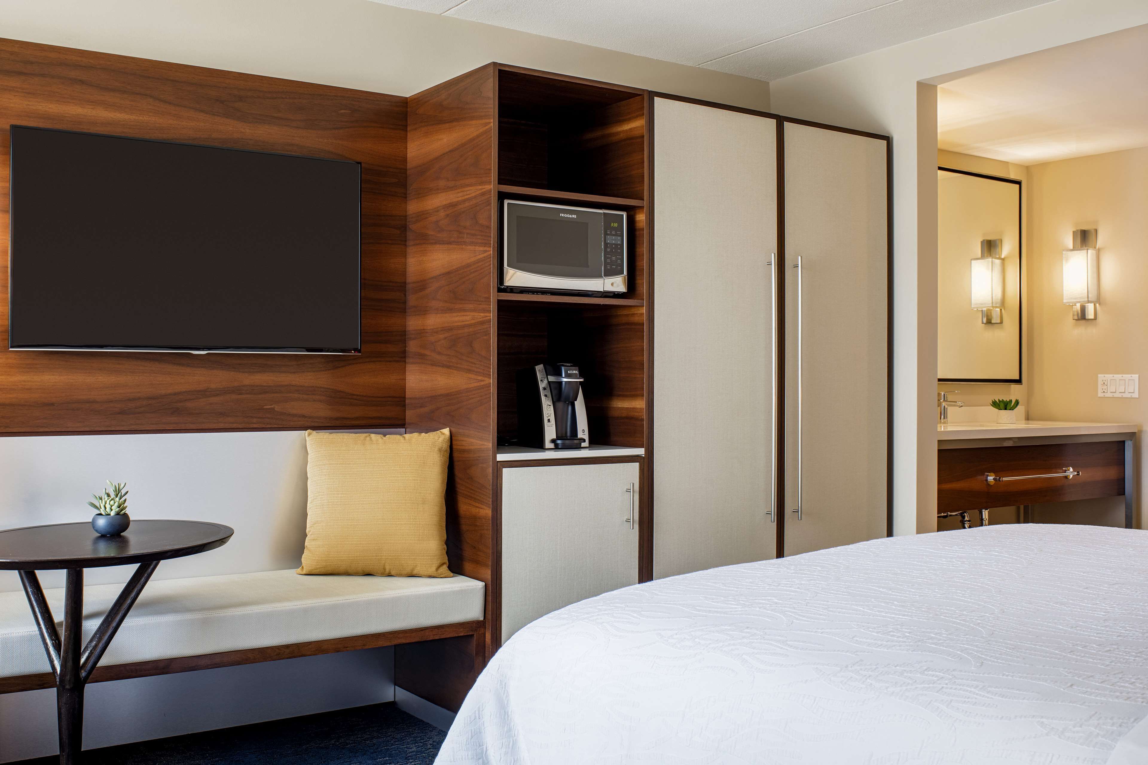 Une chambre d’hôtel comprenant un lit, un coin salon avec une table, un four à micro-ondes, un placard et une salle de bain avec un miroir et des luminaires.
