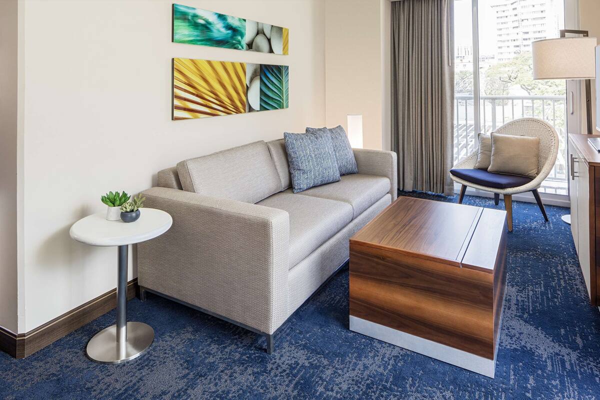 A modern hotel room with a sofa, a small round table, a chair, and a TV.