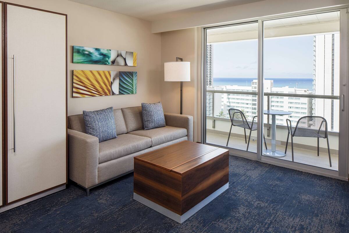 A modern hotel room with a sofa, a wooden coffee table, and a large window offering a view of the city and ocean.