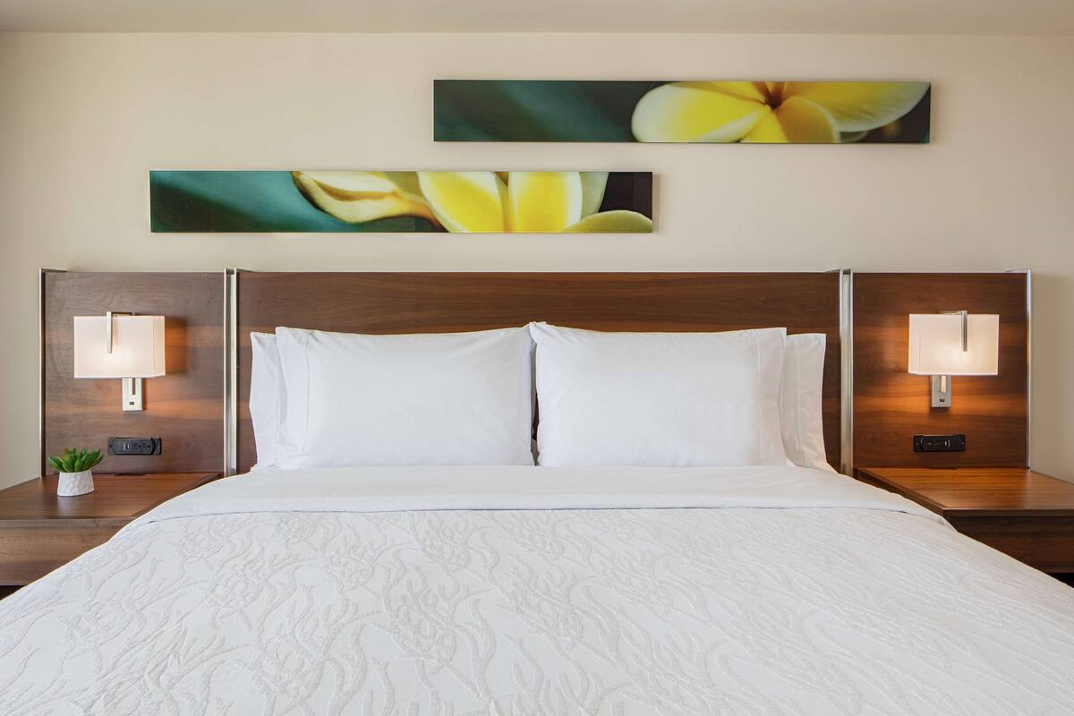A hotel room with a large bed, two wall-mounted lamps, and two framed floral paintings above the bed.