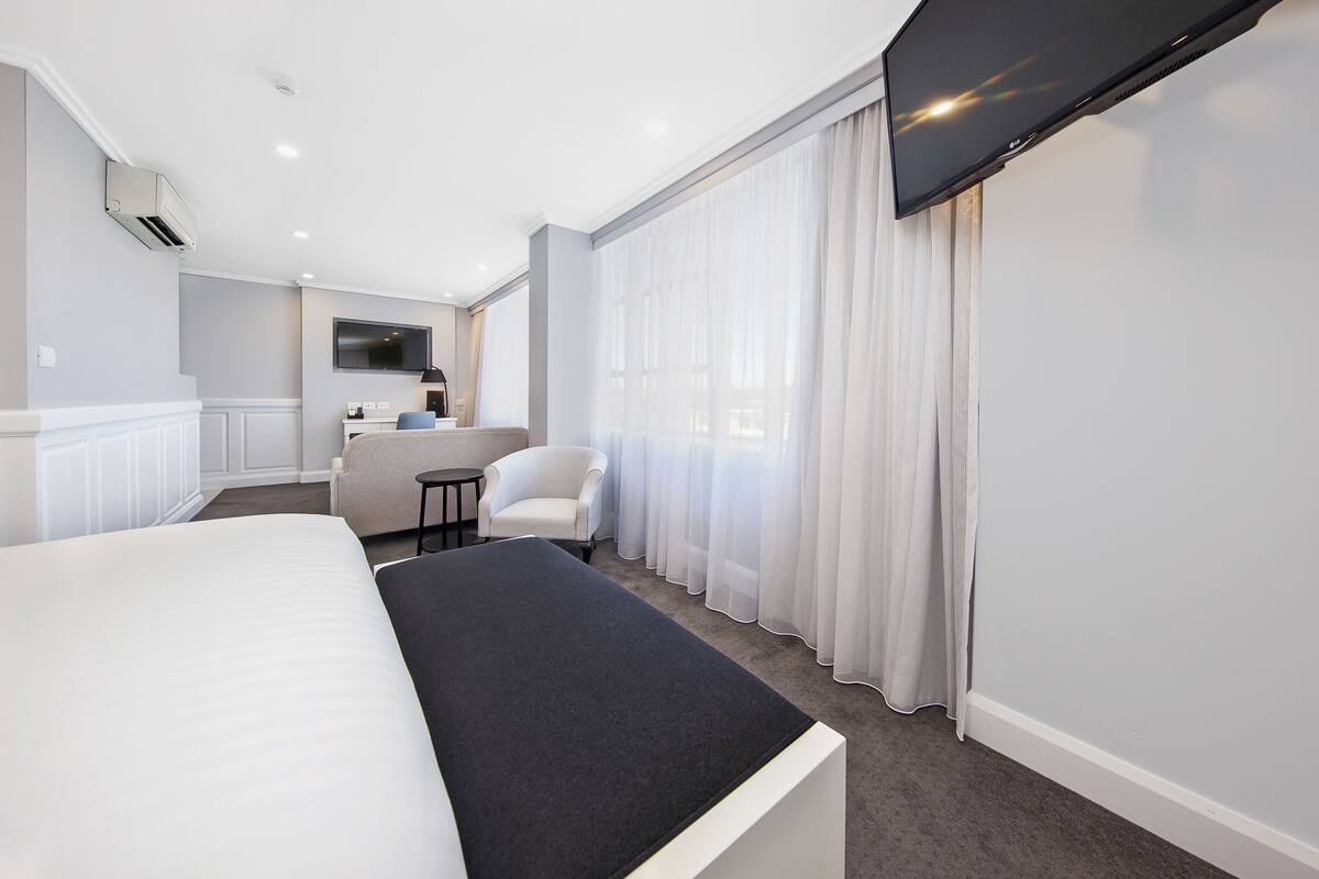 A modern hotel room with a large bed, a seating area with a chair and table, a flat-screen TV mounted on the wall, and a large window with sheer curtains.