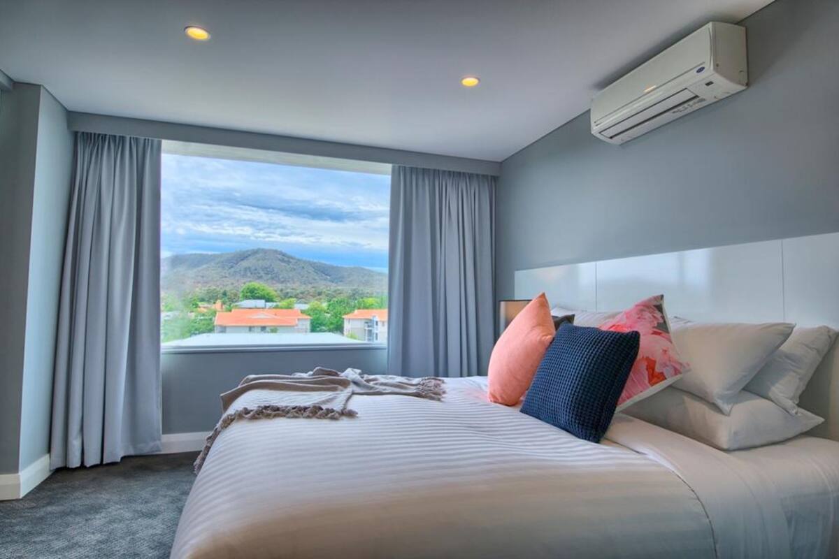 A hotel room with a large bed, grey curtains, an air conditioning unit, and a view of a mountainous landscape.